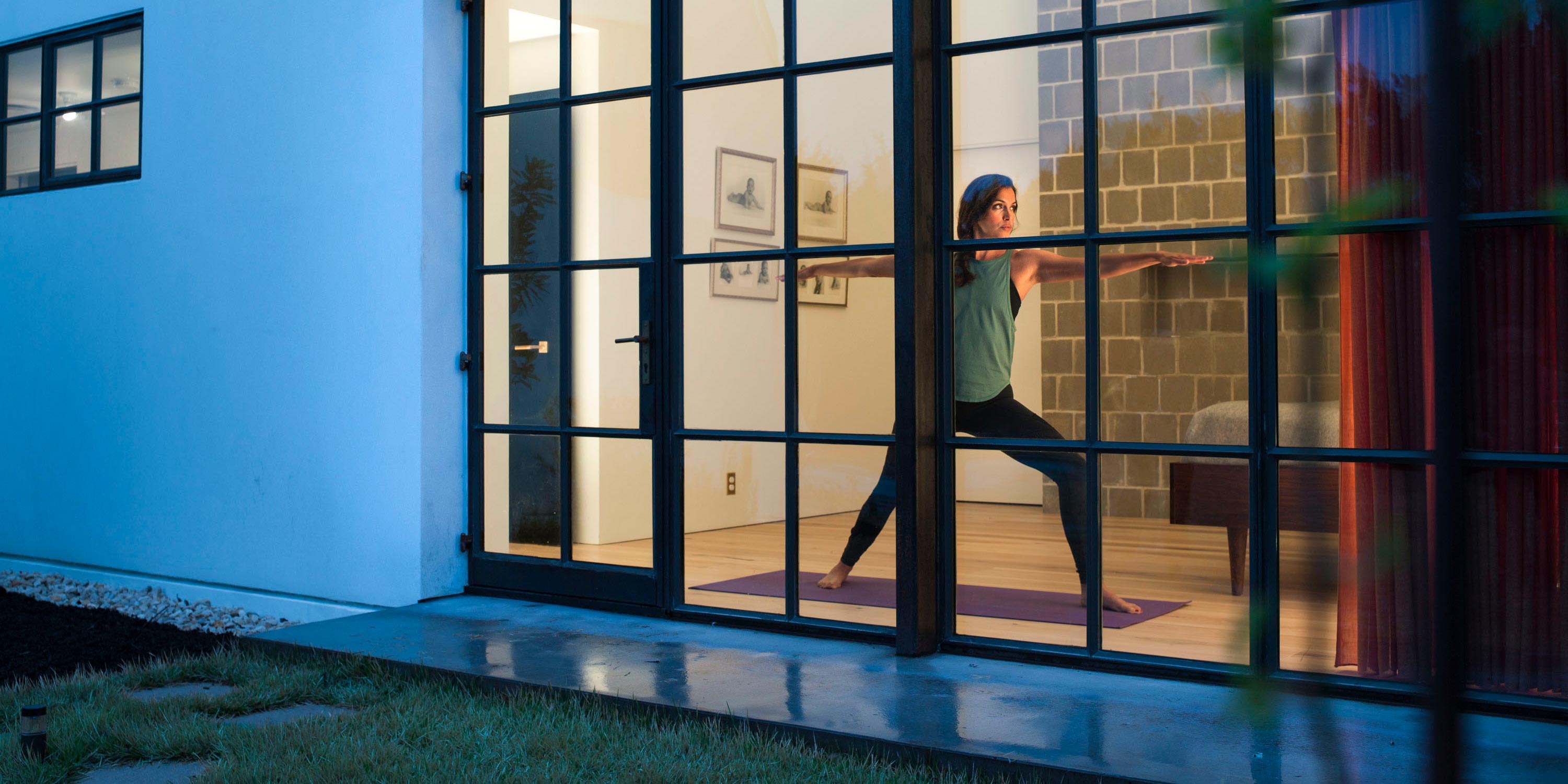 A woman practicing yoga indoors, viewed through large glass doors, performing a pose on a mat in a softly lit modern home.