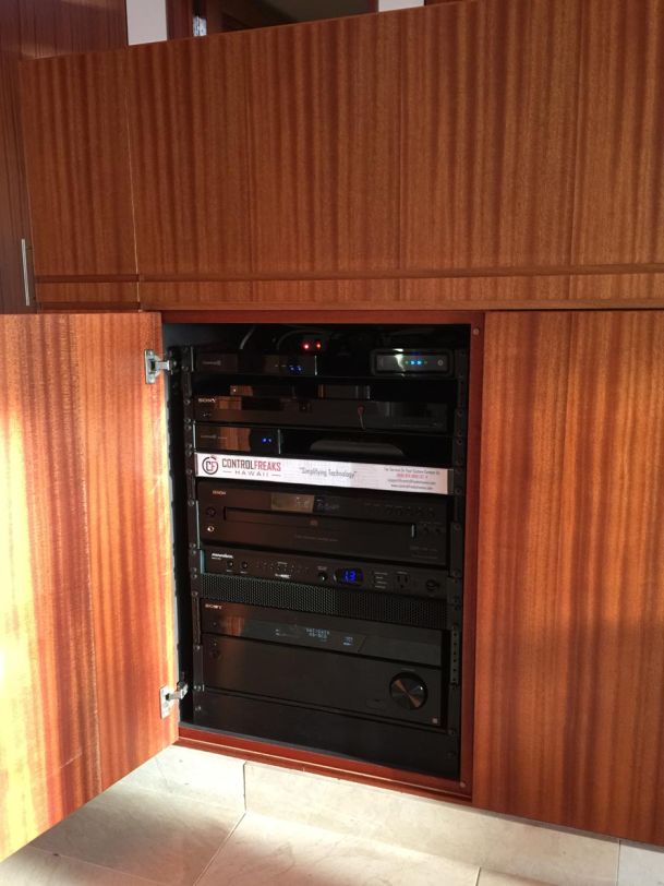 A professional audio-visual equipment rack populated with black components, including receivers and players from brands like Sony and Denon. The rack is neatly integrated into a custom wood-grain cabinet with an open door.