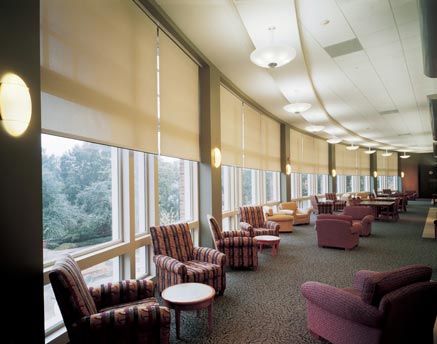 Long lounge area with a curved wall of large windows shaded by beige rollers, lined with patterned armchairs and small round tables.