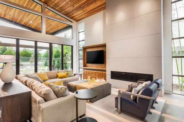 Bright living room with floor-to-ceiling windows, a wood-paneled ceiling, and a large grey sectional sofa. A flat-screen TV is mounted above a sleek, horizontal fireplace set into a tall white wall.