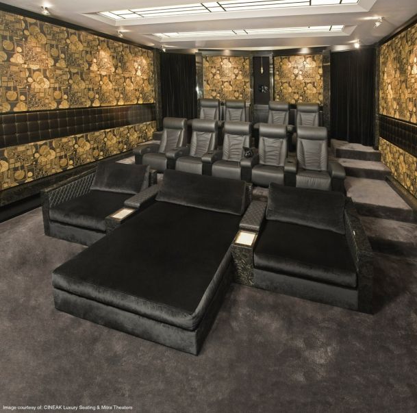 A high-end home theater featuring tiered seating with two rows of black leather recliners behind three large, plush velvet daybeds. The room is decorated with ornate gold-and-black patterned wallpaper, dark tufted wall panels, and a grey carpet. Modern recessed lighting and a sleek ceiling design complete the luxury cinema aesthetic.
